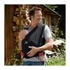 A man is holding a baby in a black sling carrier outdoors, with a wooden structure and greenery in the background. Produktbild-Vorschau