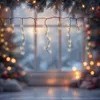 Christmas lights hanging in front of a window with a snowy scene outside, with a Christmas tree and gifts blurred in the background. Náhled obrázku produktu