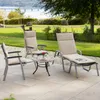 Outdoor patio furniture set with lounge chairs and a small table, set on a paved patio with a grassy background and a body of water in the distance. Produktbild-Vorschau