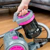 A hand is holding a detachable dustbin from a vacuum cleaner, with the main body of the vacuum cleaner visible below. The scene is set indoors on a wooden floor. Produktbild-Vorschau