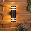A black outdoor wall light with two illuminated sections is mounted on a wooden fence. Purple flowers are visible in the foreground. Anteprima dell’immagine prodotto