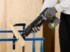 A person wearing gloves is using a reciprocating saw to cut a piece of wood, which is clamped to a blue stand. Produktbild-Vorschau