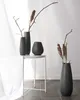 Four vases of varying sizes and shapes are arranged on and around a small white side table. Two vases are on the table, one is on the floor to the right, and the fourth is on the floor to the left, partially obscured by a curtain. Dried branches and seed p Voir les images du produit