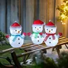 Three illuminated snowman figures with Santa hats and scarves are sitting on a wooden sled in an outdoor setting at night, with blurred lights in the background. Produktbild-Vorschau