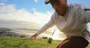 A man is in the foreground, crouching on a grassy hill overlooking the ocean. The sun is setting, casting a warm glow on the scene. Anteprima dell’immagine prodotto
