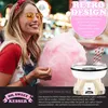 A young woman is smiling and holding a large cloud of pink cotton candy, with a retro-style cotton candy machine visible in the foreground and carnival lights in the background. Produktbild-Vorschau