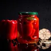 A still life composition featuring a jar of pickled red peppers, a fresh red bell pepper, garlic, and peppercorns against a dark background. Podgląd zdjęcia produktu