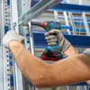 A person wearing work gloves is using a cordless drill to assemble a metal shelving unit in a warehouse or storage facility. Produktbild-Vorschau