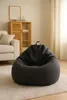 A dark gray bean bag chair sits on a light-colored rug in a living room setting. A wooden cabinet and a sofa are visible in the background. Voir les images du produit