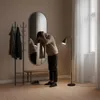 A man is adjusting his scarf in front of a mirror in a hallway with a coat rack and a floor lamp. Náhľad obrázku produktu