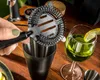 A hand is holding a cocktail strainer over a shaker, with a glass of green liquid and a lemon slice nearby. Voir les images du produit