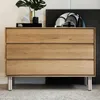 A close-up of a wooden dresser with three drawers, supported by chrome legs, placed on a textured rug. Produktbild-Vorschau