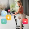 A woman is exercising on a stationary bike in a modern home setting, with various fitness tracking icons overlaid on the image. Produktbild-Vorschau