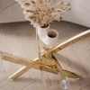 A close-up view of the base of a gold coffee table with a glass top, featuring a vase with dried flowers. Produktbild-Vorschau