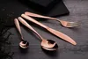 A set of rose gold cutlery is arranged on a dark wooden surface, with a stack of black napkins in the background. Produktbild-Vorschau