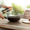 A person is spraying cooking oil into a wok on a wooden table, with a grill in the background. Produktbild-Vorschau