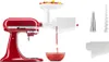 A red KitchenAid stand mixer with a white food grinder and juicer attachment is shown. The attachment is processing fruit, with juice flowing into a glass bowl below. Several separate parts of the attachment are laid out to the right of the mixer. Produktbild-Vorschau