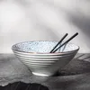 A ceramic bowl with a blue floral pattern and a brown rim, holding chopsticks, sits on a dark surface with shadows cast on a light gray and white background. Náhľad obrázku produktu