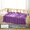 A purple fleece blanket is draped over a wooden crib, with a teddy bear and a pillow on the crib. Produktbild-Vorschau