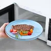 A hand holding a blue plate with a steak and vegetables in front of an open microwave. Produktbild-Vorschau
