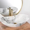 A marble basin with gold faucets is shown on a wooden surface, with a round mirror in the background. Náhled obrázku produktu