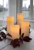 Four LED candles of varying heights are arranged on a white wooden surface, with dark red leaves and berries as decoration. The background shows a window with a blurred outdoor scene. Podgląd zdjęcia produktu
