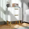 A white nightstand with gold legs and handles sits next to a bed. Decorative items are on top of the nightstand. Produktbild-Vorschau