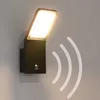 A black LED outdoor wall light with a motion sensor is mounted on a light gray wall. White wavy lines emanate from the light, indicating motion detection. Produktbild-Vorschau