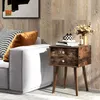 A nightstand with two drawers is placed next to a sofa in a living room setting. Náhled obrázku produktu
