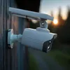 A solar-powered dummy security camera with an LED light is mounted on a wooden wall during sunset. Produktbild-Vorschau