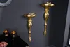 Two ornate gold wall consoles are mounted on a dark gray wall. A black table with two decorative candle holders and a small bouquet of pink flowers is visible in the foreground. Produktbild-Vorschau