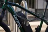 A close-up shot of a green bicycle frame with a black folding lock attached to it, parked at a bike rack. Produktbild-Vorschau