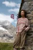 A woman leans against a stone wall in a mountainous landscape, with a flag flying in the background. Produktbild-Vorschau