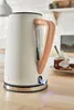 A close-up shot of a white electric kettle with a wooden handle, placed on a wooden countertop. A jar of jam and some toast are visible in the background. Podgląd zdjęcia produktu