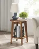 A round wooden end table with a lamp and plant on top, and books and a lantern on the shelf below. Náhled obrázku produktu