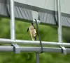 A close-up view of a ratchet strap securing a grey tarp to a silver metal frame, with a blurred green background. Podgląd zdjęcia produktu