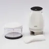 A white manual food chopper, a clear measuring cup with red markings, and a white scraper are arranged on a white background. Produktbild-Vorschau