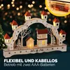 A wooden Christmas arch with LED lights, depicting houses and trees, set against a bokeh background of Christmas lights. Náhled obrázku produktu