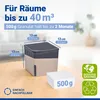A beige and gray dehumidifier sits on a wooden table next to a 500g bag of granules. The background shows a blurred interior scene with plants. Náhled obrázku produktu