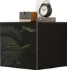 A black high-gloss nightstand with a tropical plant motif on the drawer fronts, topped with decorative items including a vintage-style alarm clock and a small amplifier. Podgląd zdjęcia produktu