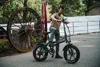 A man is standing next to a black electric bicycle in an outdoor setting with a large rusty wheel in the background. Produktbild-Vorschau
