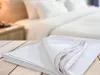 A folded white bedsheet is in the foreground on a wooden surface, with a blurred background of a white bed. Náhled obrázku produktu