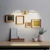 A wall-mounted light fixture above a collection of framed pictures and decorative items on a desk. Produktbild-Vorschau