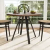 A round dining table with black metal legs and a wooden top, set with a vase of greenery and fruit in a bowl. Two chairs with woven seats and black frames are visible, along with a rug underneath the table. Anteprima dell’immagine prodotto