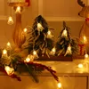 Christmas-themed decorative lights shaped like bells are draped around small artificial Christmas trees and greenery, creating a festive indoor scene. Náhled obrázku produktu