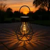 A solar garden lantern with a diamond-shaped metal frame and a bulb-shaped light is placed on a paved patio at sunset, casting intricate shadows. Produktbild-Vorschau