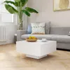 A white coffee table is placed in front of a grey sofa with cushions, next to a large potted plant. The scene is set in a living room with wooden flooring and a radiator. A framed picture hangs on the wall above the sofa. Voir les images du produit