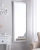 A full-length mirror with a white frame is displayed against a light gray wall. A white bedside table with a drawer is visible to the left, and a portion of a beige armchair is seen in the reflection of the mirror. Náhled obrázku produktu