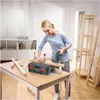A woman is using a jigsaw to cut a piece of wood on a table. There is a toolbox and other wooden pieces on the table. Shelving units are visible in the background. Anteprima dell’immagine prodotto