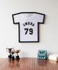 A framed t-shirt artwork hangs on a white wall above a wooden surface with trophies and a decorative box. Náhled obrázku produktu